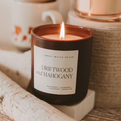 The Sweet Water Decor Driftwood and Mahogany 15 oz Candle in a matte jar sits on a wooden surface near white birch logs, with a mug and another fall candle softly blurred in the background.