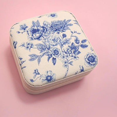 The JOSSLYN by wall to wall Blue Fleur Travel Jewelry Box, with cream vegan leather, blue floral patterns, and rounded edges, sits closed on a pink background—offering elegant accessory storage for your journeys.
