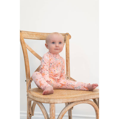 A baby wears the Sweet Bamboo Convertible Romper - Puppy Love in pink with animal patterns, sitting on a wooden chair with a woven seat against a plain white background. The soft fabric ensures comfort as the baby looks slightly upward.