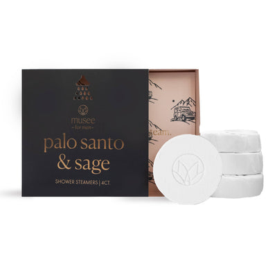 A partially open Musee Palo Santo & Sage Shower Steamers box (4 ct.) is shown with a decorative inner lid and three round white steamers infused with essential oils stacked next to it.