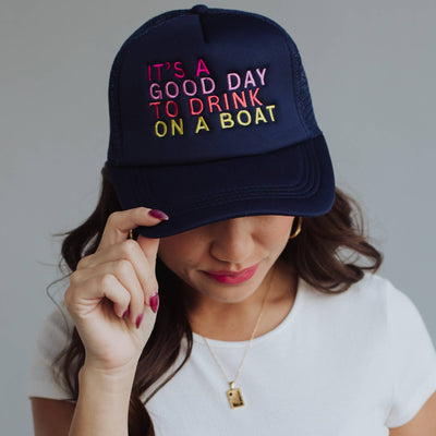 A woman wears the Navy Drink On A Boat Trucker Hat from Panache Apparel Co., featuring colorful text. Her long brown hair drapes over a white shirt as she adjusts the brim of her adjustable snapback.