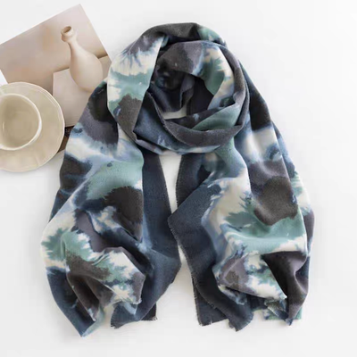 The Beaumont Scarf by Pretty Persuasions, a 72x26" 100% polyester piece in blue, green, gray, and white abstract patterns, lies beside a beige cup and saucer, white vase, and brown paper—adding elegance to any setting.