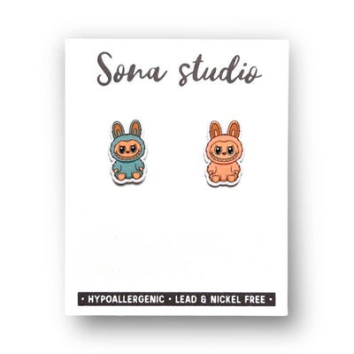 A pair of cute Little Monster Earrings—one teal, one orange—by Sona Studio, displayed on a white card. These handmade earrings feature hypoallergenic stainless steel posts and are lead and nickel free.
