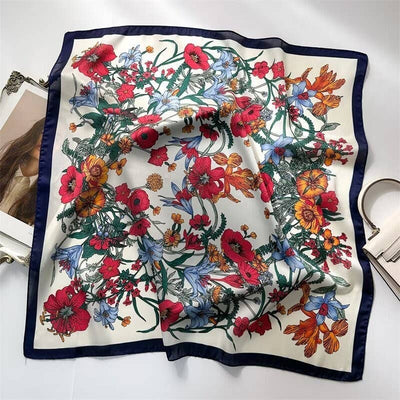 The LARGE FLORAL PRINT BANDANA SCARF by JOSSLYN by wall to wall is a square silk scarf with a dark border, showcasing vibrant red, yellow, orange, and blue flowers with green leaves on white, elegantly displayed on a white surface.