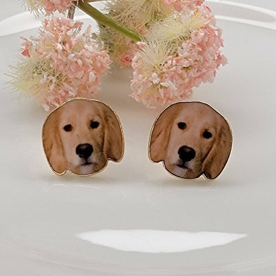 DOG PICTURE POST EARRINGS by BETTYOH INC feature golden retriever puppy faces with titanium posts, displayed on a white surface accented by pink and white floral decorations.