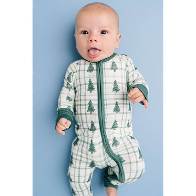 A baby lies on a light blue background wearing the Little One Shop Tree Farm Bamboo Sleeper with a white and green plaid pattern and tiny pine trees, featuring fold-over mittens, mouth open and tongue out.