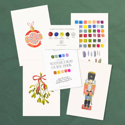 A flat lay of five holiday art cards from the Antiquaria Watercolor Painting Kit: Christmas, displayed on a green background with festive watercolor designs, a color swatch chart, and a Watercolor Guide Book card.