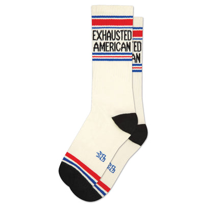 The Gumball Poodle Exhausted American Gym Crew Socks feature white fabric, black toes and heels, red and blue stripes, and bold "EXHAUSTED AMERICAN" text—ideal stress relief for any American winding down after a busy day.