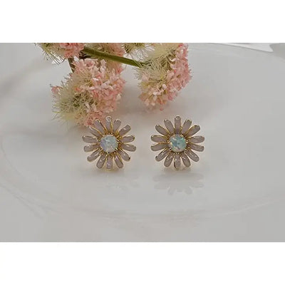 The BETTYOH INC Daisy Flower Post Earrings feature gold accents and lab-grown opal centers, displayed on a white surface with soft pink flowers blurred in the background.