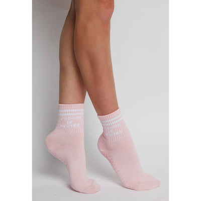 A person stands on tiptoes wearing J'adore Le Pilates Grip Socks by Pilates University 1883 in light pink with white stripes, non-slip grips, and white text near the top, set against a plain white background.