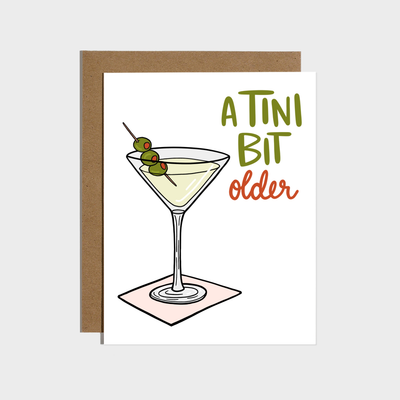 The "A Tini Bit Older Martini Birthday Card" by Brittany Paige is perfect for martini lovers, featuring a martini glass with three olives on a pink coaster and the message "A TINI BIT older" in green and orange, plus a brown envelope.