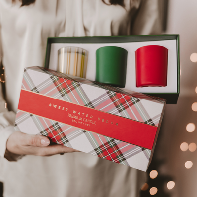 A person holds Sweet Water Decor's Soy Christmas Candle Gift Box Set—an elegant trio of candles in a plaid-patterned box. Warm, blurred holiday lights in the background evoke festive, seasonal fragrance vibes.