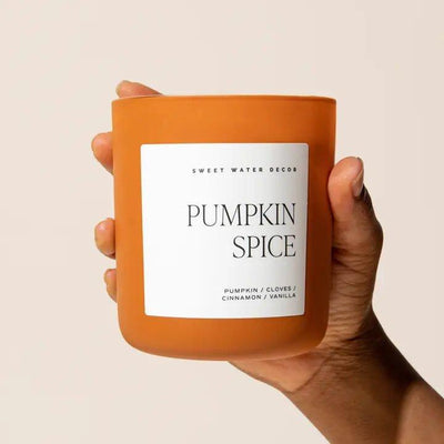 A hand holds the Sweet Water Decor Pumpkin Spice 15 oz Soy Candle in a matte jar, featuring a white label with notes of pumpkin, cloves, cinnamon, and vanilla. The background is plain light beige.
