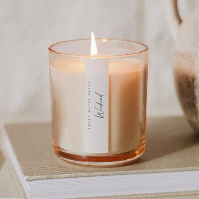 A lit 12 oz Weekend soy candle by Sweet Water Decor in a clear glass holder rests on a closed book. Blurred in the background, part of a ceramic vase is seen on the right.