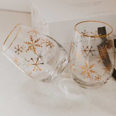 The Sweet Water Decor Snowflakes Stemless Wine Glasses, Boxed Set of 2, feature gold foil snowflake designs and gold rims. One upright glass and one on its side rest on a white surface, with the gift box in the background.