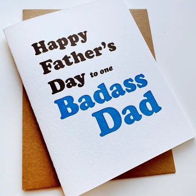 The Badass Dad Fathers Day Card by Steel Petal Press, resting on a brown envelope, is made from recycled cotton rag paper and features Happy Fathers Day to one Badass Dad in bold black and blue text, highlighting the charm of hand-printed elegance.