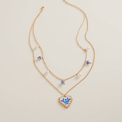 The Heart Enamel Charm Ceramic Bead 2 Layered Necklace by Designature New York features ceramic beads, small pearls, blue gemstones, and a heart-shaped pendant with a blue floral design, shown on a neutral background.