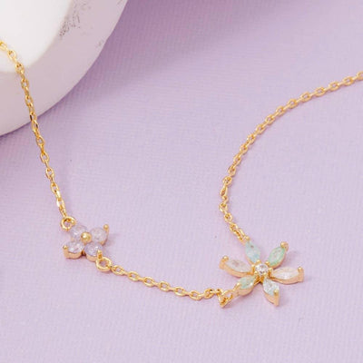 The Oberon Charm Necklace by Fame Accessories is a delicate gold-dipped piece made in Korea, featuring two floral pendants with iridescent gemstones and double flower cubic zirconia, shown on a light purple surface.