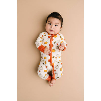 A baby with dark hair lies on a beige background, wearing the Little One Shop Pumpkin Patch Bamboo Sleeper featuring orange and yellow pumpkins and an orange zipper, looking up calmly.