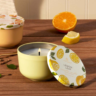 A lit Lemon Leaf Poured Candle Tin by Thymes, lemon-scented in a decorative yellow tin with lemons and leaves on the lid. A hint of lemon verbena enhances the scene with half an orange and a lemon wedge on a wooden surface.