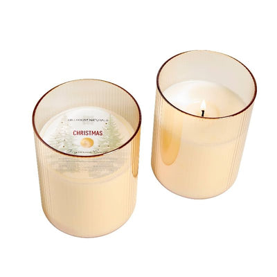 Two cream-colored, holiday-scented 15oz Christmas candles by Hillhouse Naturals/Field+Fleur in gold ribbed glass jars. One is lit, while the other features a tree image and promises long burn time for festive ambiance.