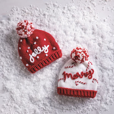 Mud Pie Holiday Knit Hats: includes a red hat with white dots, pom, and “jolly,” and a white hat with red stripes, pom, and “merry”—festive styles for 0-6 months, shown on faux snow.