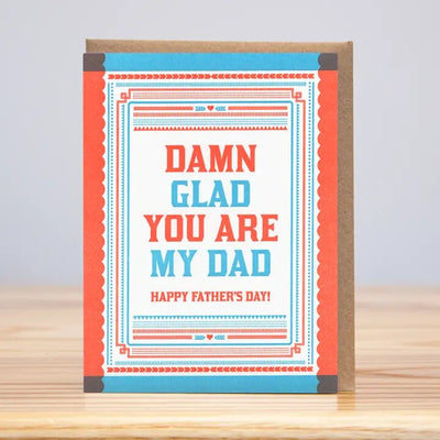 The Huckleberry Letterpress Damn Glad You Are My Dad Fathers Day card features red and blue text on quality Mohawk Paper, with a brown border. It reads, Damn glad you are my dad. Happy Fathers Day! and proudly stands upright on a wooden surface.