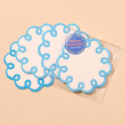 Four Coquette Scalloped Vellum Coasters - Cornflower Blue by And Here We Are, white with blue scalloped edges, are fanned on a beige surface. One remains in its clear packaging with a blue and yellow label. Set of 4.