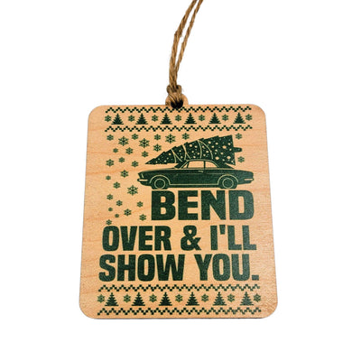 The Driftless Studios Bend Over & I'll Show You Christmas Ornament is a 3.5"x3.25" birchwood piece featuring a car, Christmas tree, snowflakes, and the phrase “BEND OVER & ILL SHOW YOU,” hanging from a brown string.
