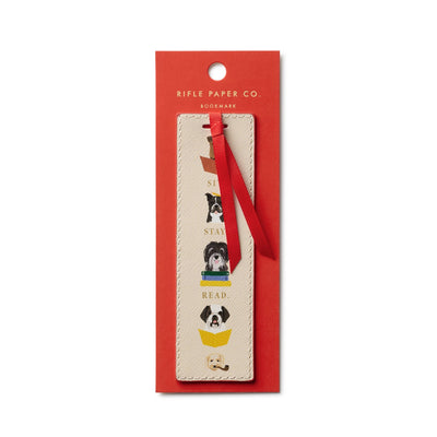 The Studious Dogs Bookmark by Rifle Paper Co. features playful dog illustrations, books, and Sit, Stay, Read on beige, attached to red packaging with a grosgrain ribbon and labeled with the brand name.