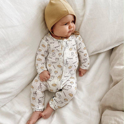 A baby in the Bellabu Bear Milk and Cookies Original Bamboo Convertible Footie sits on a soft white bed, gazing off to the side.