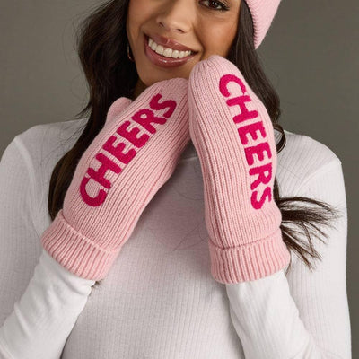 A smiling woman in a pink hat wears Panache Apparel Co. Pink Cheers Knit Mittens—fleece lined, acrylic, with bold red CHEERS lettering—posing with her mittened hands near her face and pairing them with a white long-sleeve top.