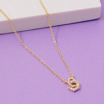 The Fame Accessories Cz Hexagonal Nuts Charm Necklace features a gold-dipped chain and a hexagon pendant with clear crystals, displayed on a purple surface beside a white rectangular object.