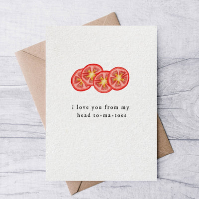 The I love from my head to-ma-toes greeting card by la nouille studio, printed on bright white paper, showcases a colorful illustration of tomato slices with playful text and comes with a kraft envelope against a light wooden background.