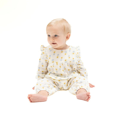 A baby with short blonde hair sits on a white background, smiling and wearing the Western Sunflower Ruffle Sleeve Romper by Angel Dear—a cream organic cotton muslin romper featuring a yellow floral pattern.