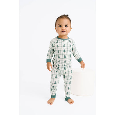 A toddler stands barefoot on a white background, wearing the Tree Farm Bamboo Pajama Set by Little One Shop. The snug pajamas feature green cuffs and Christmas tree prints. The child looks upward curiously, with one hand on a white cylindrical object.