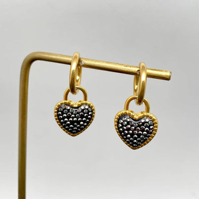 The Mio Queena Black Rhinestone Heart 18K Gold Huggie Earrings feature black and silver heart-shaped charms, shown on a gold stand against a plain white background.