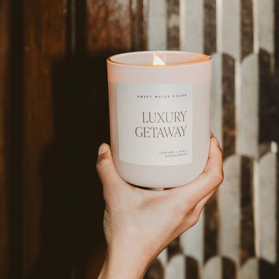 A hand holds a lit Luxury Getaway 15 oz Soy Candle by Sweet Water Decor, emanating notes of leather, amber, and sandalwood with hints of vetiver. A patterned wall provides an elegant backdrop.