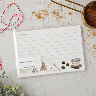 Christmas baking recipe cards by Emily Lex Studio, featuring watercolor illustrations of baked goods and utensils, are wrapped in plastic on a white surface beside a gold measuring cup with brown sugar, red-and-white twine, and greenery.