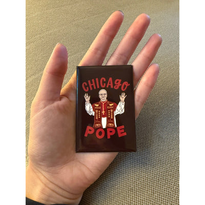 A hand holds the Lettering Works Chicago Pope Leo XIV Rectangle Magnet, featuring a vibrant illustration of the pope in red and gold robes with “CHICAGO POPE” text above and below on a dark background.