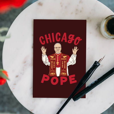 The Chicago Pope Leo XIV Postcard by Lettering Works features an illustrated pope with raised hands, CHICAGO POPE in red text above and below, shown on a marble table with a black pen and ink bottle nearby.