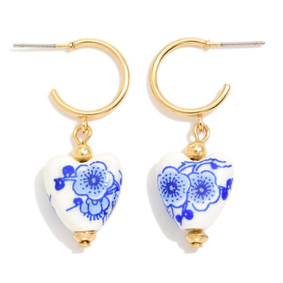 Judson & Companys Porcelain Chinoiserie Heart Earrings are gold-tone hoops (1.5 diameter) with heart-shaped pendants, featuring white porcelain adorned with blue flower patterns and accented by a small gold bead.