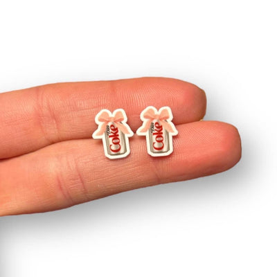 A hand displays two small Diet Soda Earrings by Sona Studio, shaped like soda bottles with bows on top. Set against a white background, these nickel free earrings are ideal for sensitive ears.