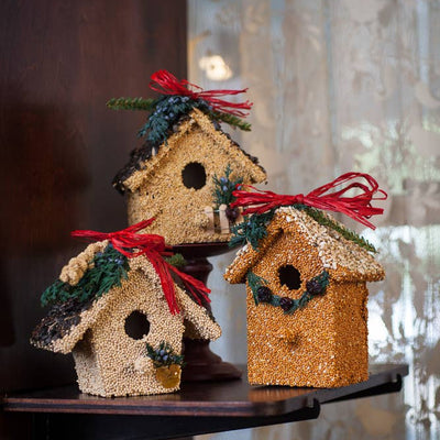 Three Holiday Birdie Cottages by Mr Bird, each adorned with seeds, greenery, and a red ribbon, create a charming gift set perfect for indoor display.