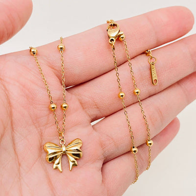 A hand holds the Mio Queena Gold Plated Stainless Steel Bow Pendant Necklace, featuring a bow-shaped charm, small round beads along the chain, a lobster clasp, and a branded tag near the clasp.