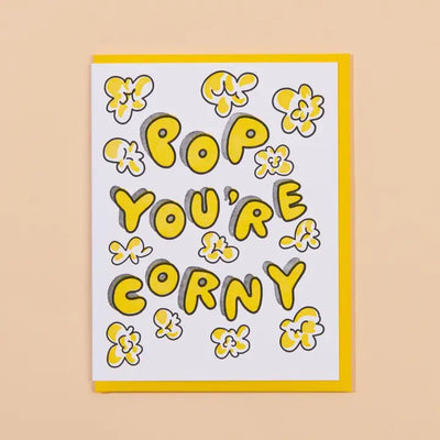 The Corny Pop Fathers Day Greeting Card by And Here We Are features the text POP YOURE CORNY in bold yellow letters outlined in black, surrounded by cartoon popcorn illustrations. Its letterpress printed on cotton paper with an A2 fold and a yellow border on a beige background.