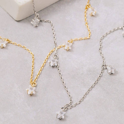 The Fame Accessories CZ Star Station Necklace set features two dainty chains—one gold-dipped cubic zirconia and one silver—each adorned with small CZ star charms, beautifully displayed on a light grey surface.