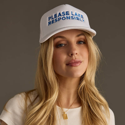 A person wears a Panache Apparel Co. Please Lake Responsibly embroidered white trucker hat, along with a white shirt and rectangular pendant necklace, against a plain background.