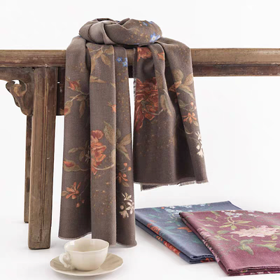 The Pretty Persuasions Fall Forest Scarf in brown floral polyester (74"x27") is draped on a rustic table. Nearby, a teacup sits next to two folded Fall Forest Scarves in purple and blue on a white surface.