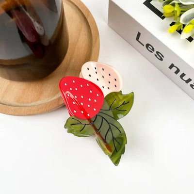 A 7.5cm acetate Strawberry Pair Hair Claw Clip by The Diva Soap, shaped like a strawberry with green leaves and a small beige section with black dots, rests on wood beside a dark beverage in a glass and a white box with partial Les nuages text.